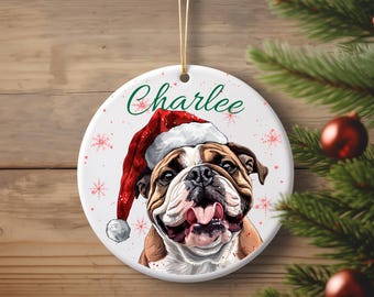 May include: A round Christmas ornament featuring a bulldog wearing a Santa hat. The ornament has the name "Charlee" in green script. Red snowflakes are scattered around the image of the dog. The ornament hangs from a gold string.