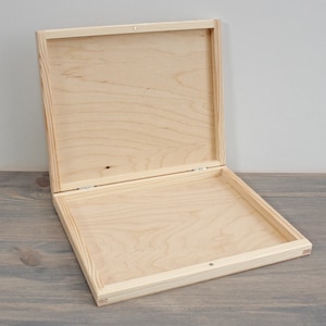 Könnte beinhalten: Eine rechteckige Holzkiste mit aufklappbarem Deckel. Die Kiste ist aus hellem, unbehandeltem Holz gefertigt, mit sichtbarer Maserung. Der Deckel ist leicht geöffnet und gibt den Blick ins Innere frei. Die Kiste steht auf einer Holzoberfläche.