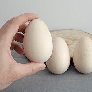 Puede incluir: Tres huevos de madera sin terminar, uno sostenido en una mano, los otros dos sobre una superficie de madera.