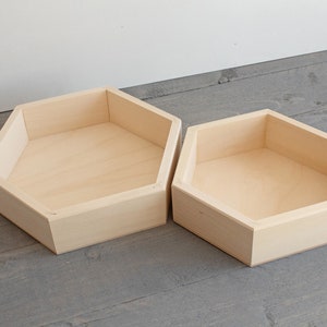 May include: Two unfinished, light-colored wooden hexagon-shaped trays. The trays have a simple, geometric design with angled sides and a flat base. The wood grain is visible, and the trays appear to be empty. They are likely used for storage or display.