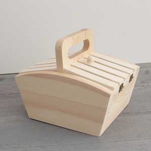 May include: A light-colored wooden box with a handle and a slatted lid. The box has a rectangular shape with curved sides and is secured with small metal hinges. The handle is arched, and a small wooden knob is on the lid.