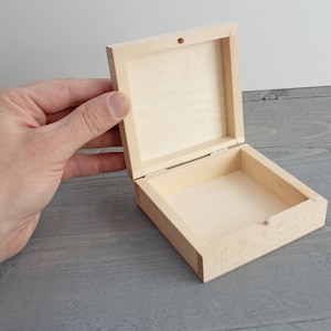 May include: A small, open, square wooden box with a hinged lid. The box is made of light-colored wood and has a simple, minimalist design. The lid is slightly ajar, revealing the empty interior. The box is held by a hand.