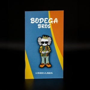 May include: A yellow, blue, and black enamel pin featuring a cartoon skull wearing a gold crown and a yellow and blue patterned jacket. The pin is on a card with the text "Bodega Bros" in black and white.