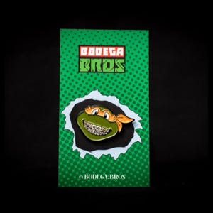 Könnte beinhalten: Grün-orangefarbene Teenage Mutant Ninja Turtles-Anstecknadel mit grünem Gesicht, orangefarbenem Bandana und silbernen Zahnspangen. Die Anstecknadel befindet sich auf einem grün-weißen Hintergrund mit dem Text "Bodega Bros" in Rot und Schwarz.