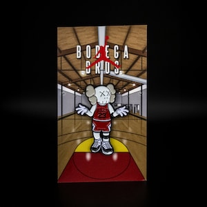 May include: A white and red enamel pin featuring a cartoon character wearing a red and white basketball jersey with the text "Bulls 23". The character is standing on a red basketball court with the text "Bodega Bros" above.