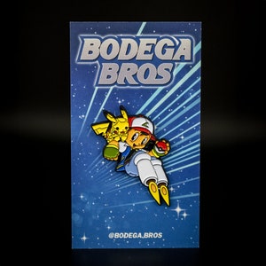 May include: A blue and white enamel pin featuring a cartoon character from the Pokemon franchise. The character is wearing a red and white hat and holding a yellow Pokemon character. The pin has the text "BODEGA BROS" on the bottom.