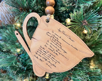 May include: A wooden ornament shaped like a mug with a handwritten recipe for "Hot Fruit Salad" on it. The recipe includes ingredients like coconut macaroons, mandarin oranges, and brown sugar. The ornament is hanging on a Christmas tree.