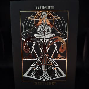 May include: A black tarot card box with gold foil accents. The box features the text "INA AUDERIETH" at the top and "CONJUNCTION TAROT" at the bottom. The artwork depicts a figure in meditation, surrounded by geometric and celestial designs.