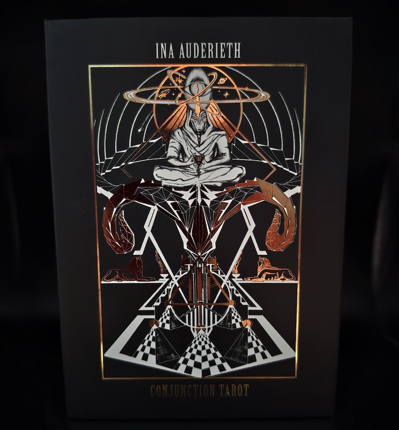 May include: A black tarot card box with gold foil accents. The box features the text "INA AUDERIETH" at the top and "CONJUNCTION TAROT" at the bottom. The artwork depicts a figure in meditation, surrounded by geometric and celestial designs.