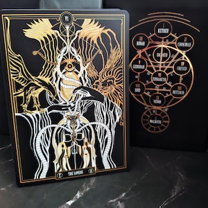 May include: A black tarot card with gold and white illustrations. The card features the text "THE LOVERS" and the Roman numeral "VI". The design includes winged figures, snakes, and a chalice. A second card with a gold Kabbalistic Tree of Life is also visible.