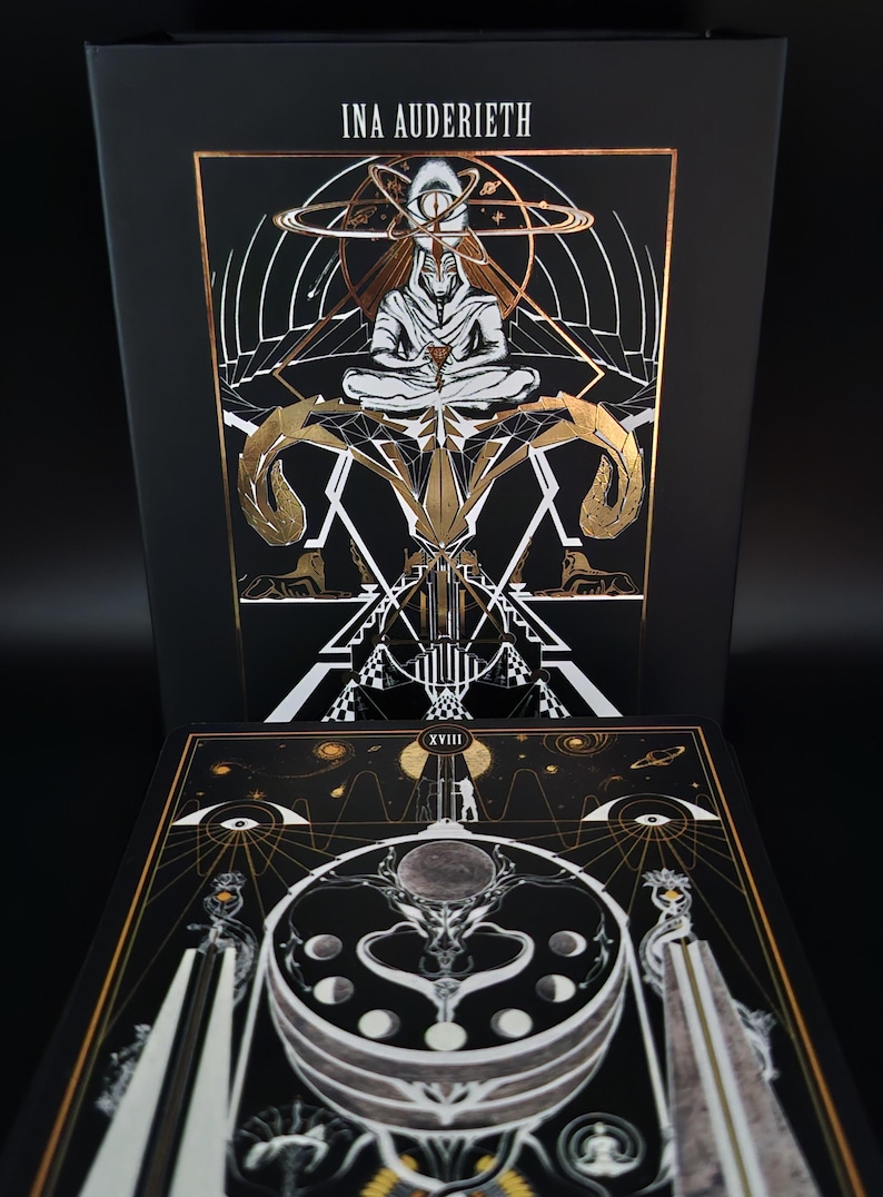 May include: Black tarot card box with gold foil accents and the name "INA AUDERIETH" at the top. The box features an illustration of a figure in meditation, surrounded by celestial elements. The bottom card displays a celestial design with the Roman numeral XVIII.