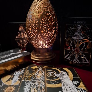 May include: A gold-colored, teardrop-shaped lamp with intricate cut-out patterns, illuminating a warm glow. Tarot cards with black and gold illustrations are displayed, along with a tarot card box. The cards feature detailed artwork and the text "CONJUNCTION TAROT".