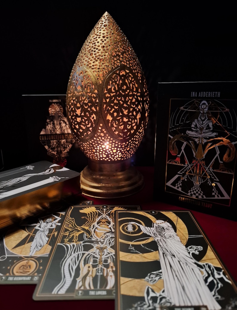 May include: A gold-colored, teardrop-shaped lamp with intricate cut-out patterns, illuminating a warm glow. Tarot cards with black and gold illustrations are displayed, along with a tarot card box. The cards feature detailed artwork and the text "CONJUNCTION TAROT".
