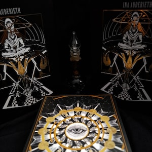 May include: Two rectangular black books titled INA AUDERIETH featuring a white and gold illustration of a seated figure, and a central square book with a gold and white eye mandala design, surrounding a small clear glass bottle with a pointed stopper.