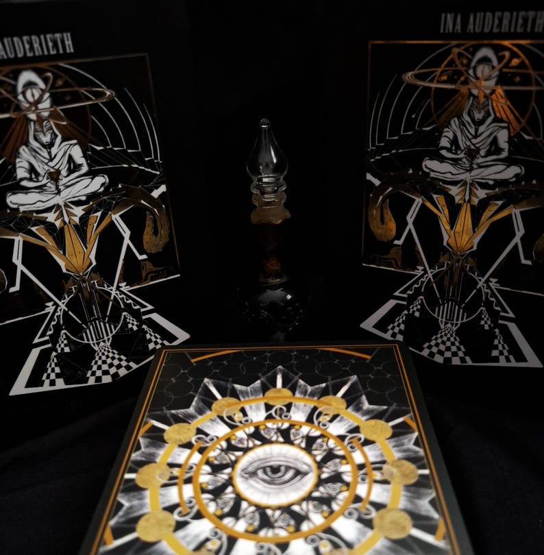 May include: Two rectangular black books titled INA AUDERIETH featuring a white and gold illustration of a seated figure, and a central square book with a gold and white eye mandala design, surrounding a small clear glass bottle with a pointed stopper.