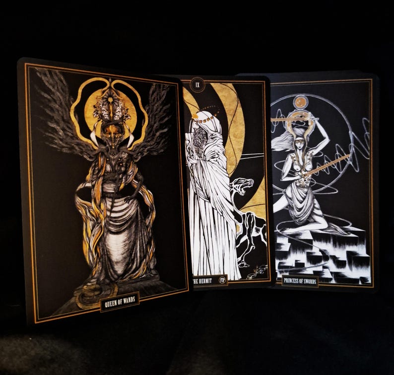 May include: Three tarot cards with black backgrounds and gold and white illustrations. The cards are labeled "Queen of Wands", "The Hermit", and "Princess of Swords". The cards have gold borders and are displayed on a dark surface.