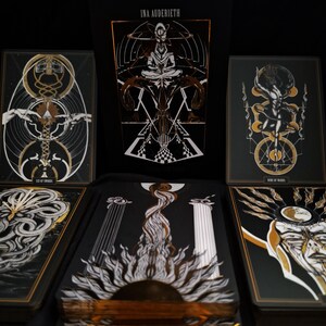 May include: A tarot deck with a black and gold color scheme. The deck features intricate illustrations with geometric and symbolic designs. The box displays the name "INA AUDERIEH". The cards are arranged to showcase the artwork.
