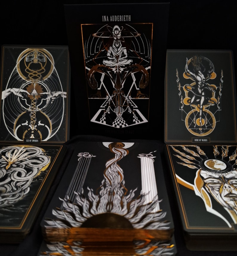 May include: A tarot deck with a black and gold color scheme. The deck features intricate illustrations with geometric and symbolic designs. The box displays the name "INA AUDERIEH". The cards are arranged to showcase the artwork.
