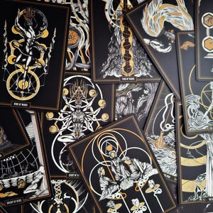 May include: A collection of tarot cards with black backgrounds and gold and white illustrations. Each card features unique artwork, including figures, symbols, and text. The cards are arranged in a scattered pattern, showcasing the detailed designs.