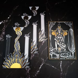 May include: A tarot card deck with a black background and gold and silver accents. The cards feature illustrations of pillars, suns, and a seated figure labeled "Queen of Disks". The deck has a mystical theme.