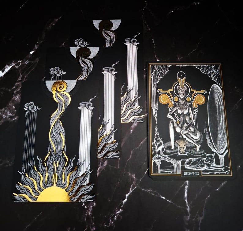 May include: A tarot card deck with a black background and gold and silver accents. The cards feature illustrations of pillars, suns, and a seated figure labeled "Queen of Disks". The deck has a mystical theme.