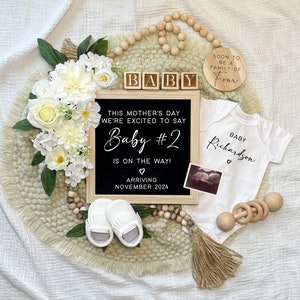 Peut inclure: Annonce de grossesse avec un tableau noir indiquant "Bébé #2 arrive en novembre 2024". L'ensemble comprend un body blanc, des chaussures de bébé, des blocs de bois épelant "BABY" et une échographie. Décoré de fleurs blanches et de perles en bois.