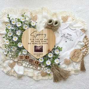 May include: A wooden heart-shaped sign with the text "Spring is in the air and we have some big news to share" and a small ultrasound image. The sign is surrounded by white daisies, wooden beads, and a white onesie with the text "Baby Richardson" and a small heart.