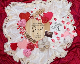 Könnte beinhalten: Ein herzförmiges Schild aus Holz mit dem Text "We've been keeping a little secret" und "Arriving July 2024". Das Schild ist umgeben von roten und rosa Herzen, einem weißen Baby-Strampler mit roten und rosa Herzen und einer weißen Decke mit Fransen.