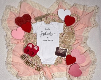 Könnte beinhalten: Eine rosa-weiße Decke mit einem weißen Strampler, auf dem "Baby Richardson Juni 2024" steht, und einem Holzschild mit der Aufschrift "Unser kleiner Valentinstag". Außerdem sind rote und rosa Herzen, ein Paar rote Babyschuhe und ein Holzblock mit der Aufschrift "Baby" zu sehen.