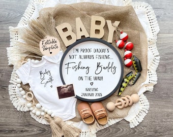 Op de afbeelding: Een zwart-wit bord met de tekst "I'm proof Daddy's not always fishing... Fishing Buddy on the way! Arriving January 2025" omringd door vislokken, een houten babyratel en een witte romper met de tekst "Baby Smith" erop.