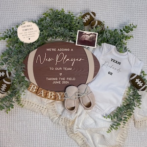 May include: A wooden football-shaped sign with the text "We're adding a New Player to our team. Taking the field June 2024."  A white onesie with the text "Team Richards 02" is next to the sign.  There are brown and white wooden blocks spelling out "BABY" and a white blanket with fringe.