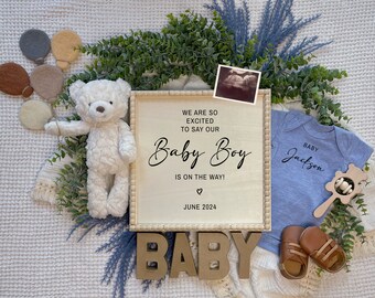 May include: A wooden sign with the text "We are so excited to say our Baby Boy is on the way! June 2024" in black lettering. A blue baby onesie with the text "Baby Jackson" in black lettering. A white teddy bear and a wooden rattle.