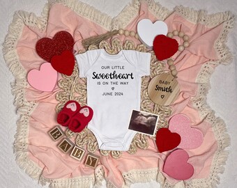 Könnte beinhalten: Eine rosa Decke mit weißen Fransen und ein weißer Strampler mit dem Text "Our little sweetheart is on the way June 2024". Es gibt rote und rosa Herzen, ein Holzschild mit der Aufschrift "Baby Smith" und einen Holzblock mit der Aufschrift "Baby".