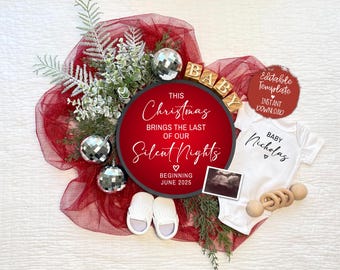 May include: A white onesie with the text "Baby Nicholas" and a heart. A round wooden sign with the text "This Christmas brings the last of our Silent Nights" and a heart. The sign has a white background and black text. The onesie and sign are surrounded by red tulle and white greenery.