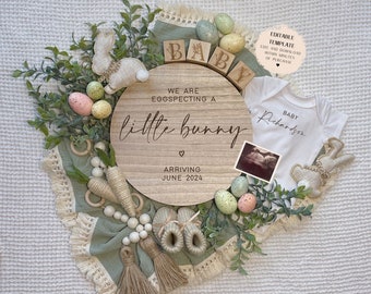 May include: A wooden round sign with the text "We are eggspecting a little bunny" and "Arriving June 2024". The sign is surrounded by green leaves, pastel colored eggs, and a white onesie with the text "Baby Richardson".