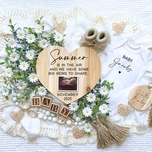 May include: A wooden heart-shaped sign with the text "Summer is in the air and we have some big news to share... November 2025". A small ultrasound image is in the center of the sign. The sign is surrounded by white daisies, wooden blocks spelling "BABY", a white onesie with the text "Baby Jacobs", a wooden round sign with the text "Hello Baby!" and other decorative items.
