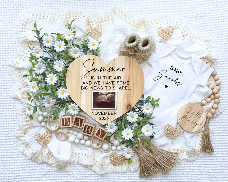 May include: A wooden heart-shaped sign with the text "Summer is in the air and we have some big news to share... November 2025". A small ultrasound image is in the center of the sign. The sign is surrounded by white daisies, wooden blocks spelling "BABY", a white onesie with the text "Baby Jacobs", a wooden round sign with the text "Hello Baby!" and other decorative items.
