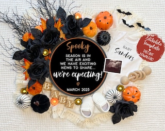May include: A Halloween-themed pregnancy announcement with a black and orange color scheme. A round sign reads "Spooky Season is in the air and we have exciting news to share... We're expecting!" with a heart and the date "March 2025". A white onesie with the text "Baby Sanders" is visible. Other decorations include black roses, pumpkins, and a wooden rattle.