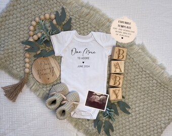 May include: A white onesie with the text "One More To Adore" and the date "June 2024".  A wooden sign reads "Our Growing Family".  Wooden blocks spell out "BABY".  The onesie, sign, and blocks are on a light green fuzzy blanket.