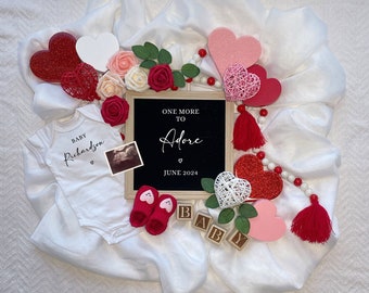 Könnte beinhalten: Ein weißer Strampler mit dem Text "Baby Richardson" und einem Herz. Ein schwarzes Schild mit dem Text "One More To Adore" und dem Datum "June 2024". Rote, rosa und weiße Herzen, eine weiße Perlenkette und rote Quasten umgeben den Strampler und das Schild.