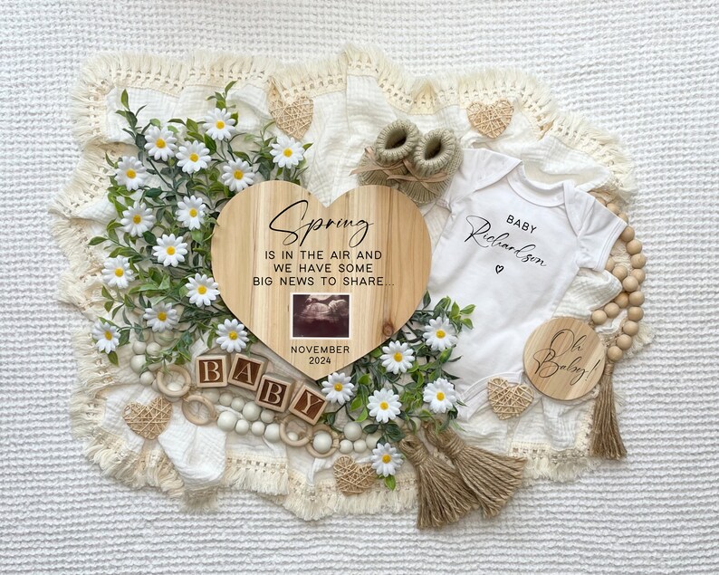 May include: A wooden heart-shaped sign with the text "Spring is in the air and we have some big news to share" and a small ultrasound image. The sign is surrounded by white daisies, wooden beads, and a white onesie with the text "Baby Richardson" and a small heart.