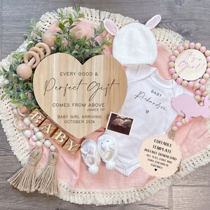 May include: A wooden heart-shaped sign with the text "Every good & Perfect Gift comes from above James 1:17 Baby Girl Arriving October 2024". A pink and white baby onesie with the text "Baby Richardson" and a small heart. A pink and white knitted baby hat. A pink wooden bunny and a wooden bead garland with pink and white beads. A set of wooden blocks spelling "BABY".