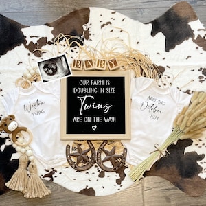 Könnte beinhalten: Ein weißer Kuhfellteppich mit zwei weißen Stramplern, einer mit dem Text "Weston Twins" und der andere mit dem Text "Arriving October 2024". Es gibt Holzblöcke, die "BABY" buchstabieren, ein Schild aus schwarzem Schiefer, auf dem steht "Our farm is doubling in size Twins are on the way!" und eine braune Holzspielzeugkuh mit einem Rassel.
