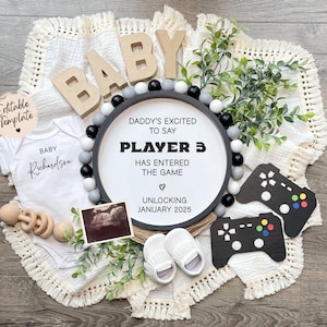 May include: A black and white baby announcement sign with the text "Daddy's excited to say Player 3 has entered the game. Unlocking January 2025". The sign is surrounded by a white blanket, a baby onesie, a rattle, a pair of booties, and a sonogram photo.