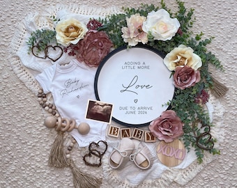 Könnte beinhalten: Ein weißer Strampler mit dem Text "Baby Richardson" und einem Herz. Ein Holzschild mit dem Text "Adding a little more Love" und "Due to arrive June 2024". Das Schild ist von Grünpflanzen und Blumen umgeben.