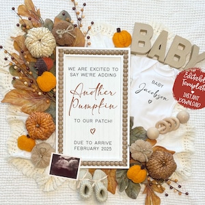 以下が含まれることがあります： 縁に茶色のビーズが付いた白い木製の看板に、「もう1つのカボチャ」が2025年2月に到着することが発表されています。看板は、カボチャ、葉、ベビーロンパースなど、秋の装飾で囲まれています。