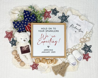 May include: A white sign with red text that reads "Hold on to your sparklers We're Expecting! Arriving January 2025". The sign is surrounded by a white teddy bear, a sonogram, a baby onesie, wooden blocks that spell "BABY", and red, white, and blue star decorations.