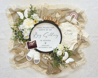 May include: A black and white sign with the text "Hand picked for earth by my Big Sibling in Heaven arriving November 2024". The sign is surrounded by white flowers, a wooden baby block spelling "BABY", a white onesie with the text "BABY Smith", a wooden beaded garland, and a white butterfly.