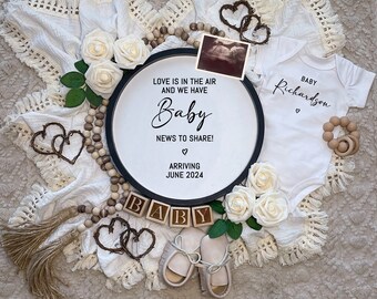 Könnte beinhalten: Ein weißer Strampler mit dem Text "Baby Richardson" und einem kleinen Herz. Eine Holzperlenkette, ein Holz-Beißring für Babys und ein rundes Holzschild mit dem Text "Love is in the air and we have Baby News to share! Arriving June 2024" sind ebenfalls auf dem Bild zu sehen.