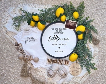 May include: A white baby onesie with the text "Baby Richardson" on it. A wooden sign with the text "We are excited to say our sweet little one is on the way! May 2024" is surrounded by lemons and greenery.  Wooden blocks spell out "BABY" and a small wooden sign says "Our Main Squeeze".